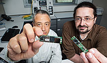 IPOS developers Luigi Gentile Polese and Larry Brackney sit at a building automation control network while showing the small size of the essential parts of the IPOS – the microprocessor and the camera. Credit: Dennis Schroeder